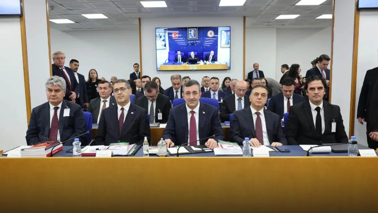 TBMM Plan ve Bütçe Komisyonu'nda kürsü masasında oturan Cevdet Yılmaz ve yanındaki bürokratlar; arkalarında ayakta duran ve oturan milletvekilleri, duvarda Genel Kurul yayını açık bir ekran.