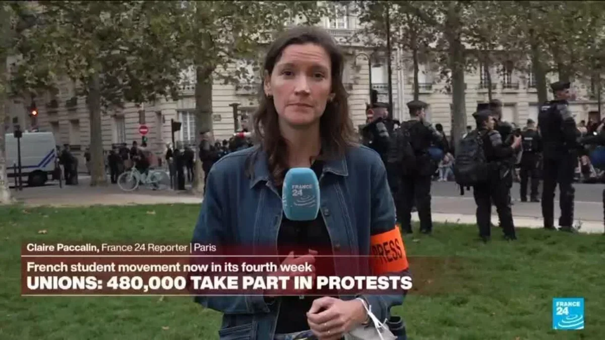 France 24 muhabiri Claire Paccalin Paris'te öğrenci eylemleri sırasında rapor veriyor; arkada taktik giyimli polisler.